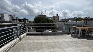 Une terrasse sur le toit, dotée d’une rambarde en métal, d’une table et de chaises, et offrant une vue sur les bâtiments et les arbres sous un ciel partiellement nuageux.