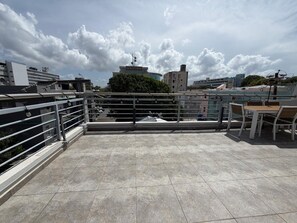 Une terrasse sur le toit, dotée d’une rambarde en métal, d’une table et de chaises, et offrant une vue sur les bâtiments et les arbres sous un ciel partiellement nuageux.