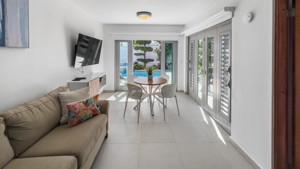 A modern living room with a sofa, a coffee table, and a TV. There is a dining area with chairs and a table. Large windows provide a view of the outdoors. - Spacious and Modern Villa with Pool in Ocean Park (San Juan)