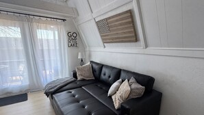 A modern living room with a black leather sofa, a wooden wall decor, and a window with sheer curtains. - Kayak Cottage - Free kayaks and fire pit (Lampe)
