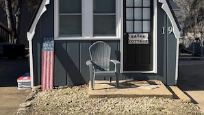 A small, dark-colored cottage with a triangular roof, a front door, and a window. A chair is placed outside, and the number 19 is visible on the building. - Kayak Cottage - Free kayaks and fire pit (Lampe)