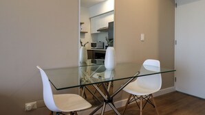 A modern dining area with a glass table and white chairs, a kitchen in the background, and a wooden floor.