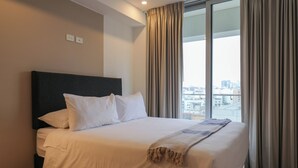 A hotel room with a large bed, a dark headboard, white bedding, a throw blanket, and a view of a cityscape through large windows with curtains.