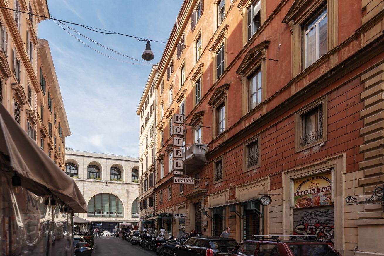 Une rue étroite, avec un hôtel appelé « HÔTEL LUCIANI » sur le côté droit, une boulangerie sur le côté gauche et des voitures garées le long de la rue.
