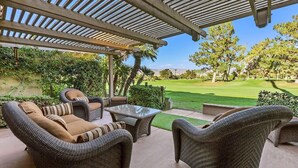 Un patio couvert aménagé avec des meubles en osier, une table à plateau en verre et une vue sur une pelouse et des arbres bien entretenus.