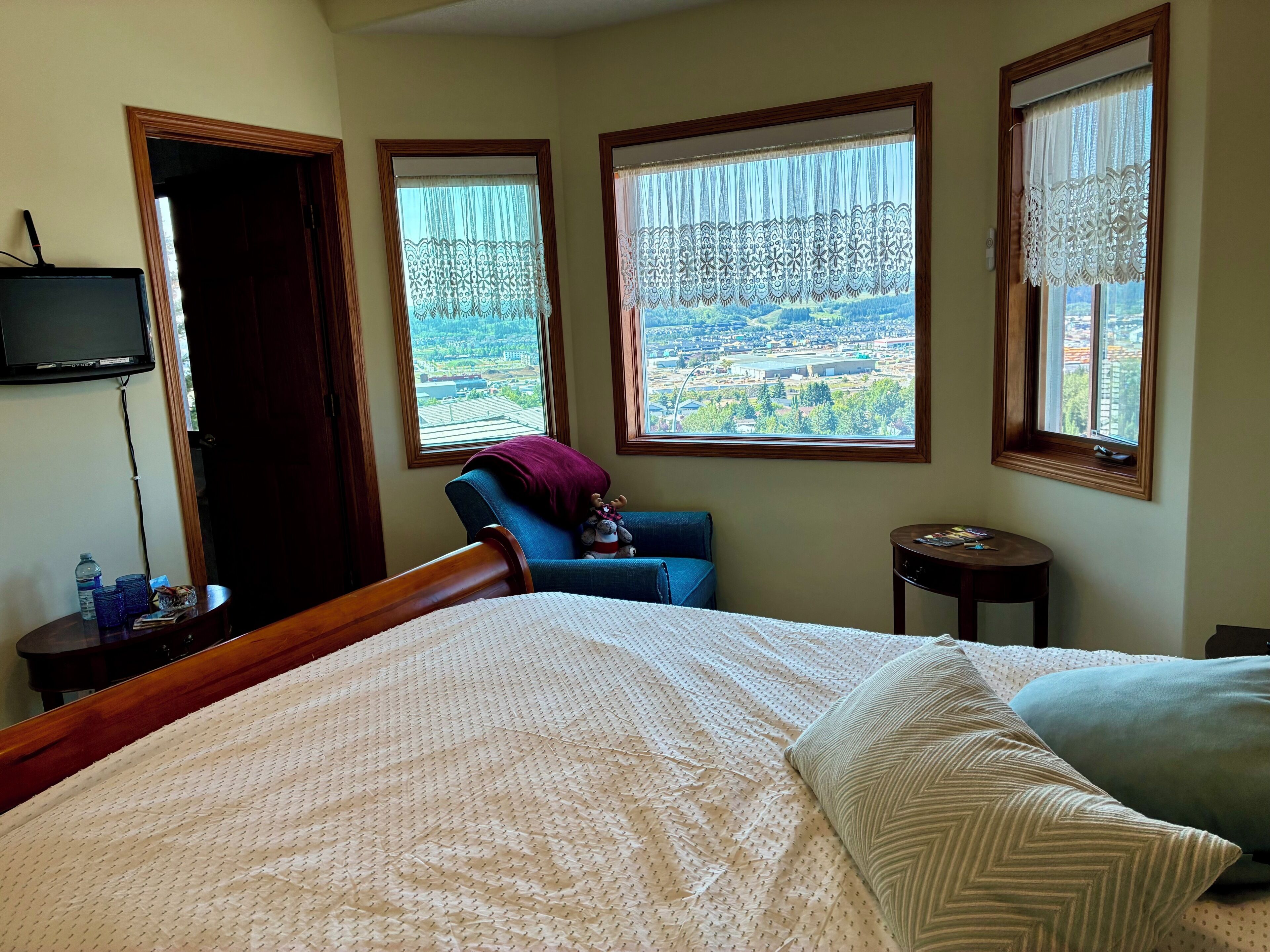Un dormitorio con una cama, una silla, una mesita y un televisor. Hay tres ventanas con cortinas.