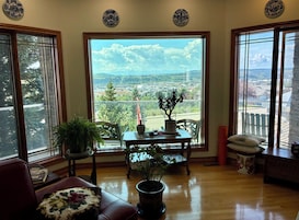 Una habitación con un ventanal amplio que ofrece vistas a la ciudad, una mesa de madera con planta en maceta y una zona de estar con cojines.