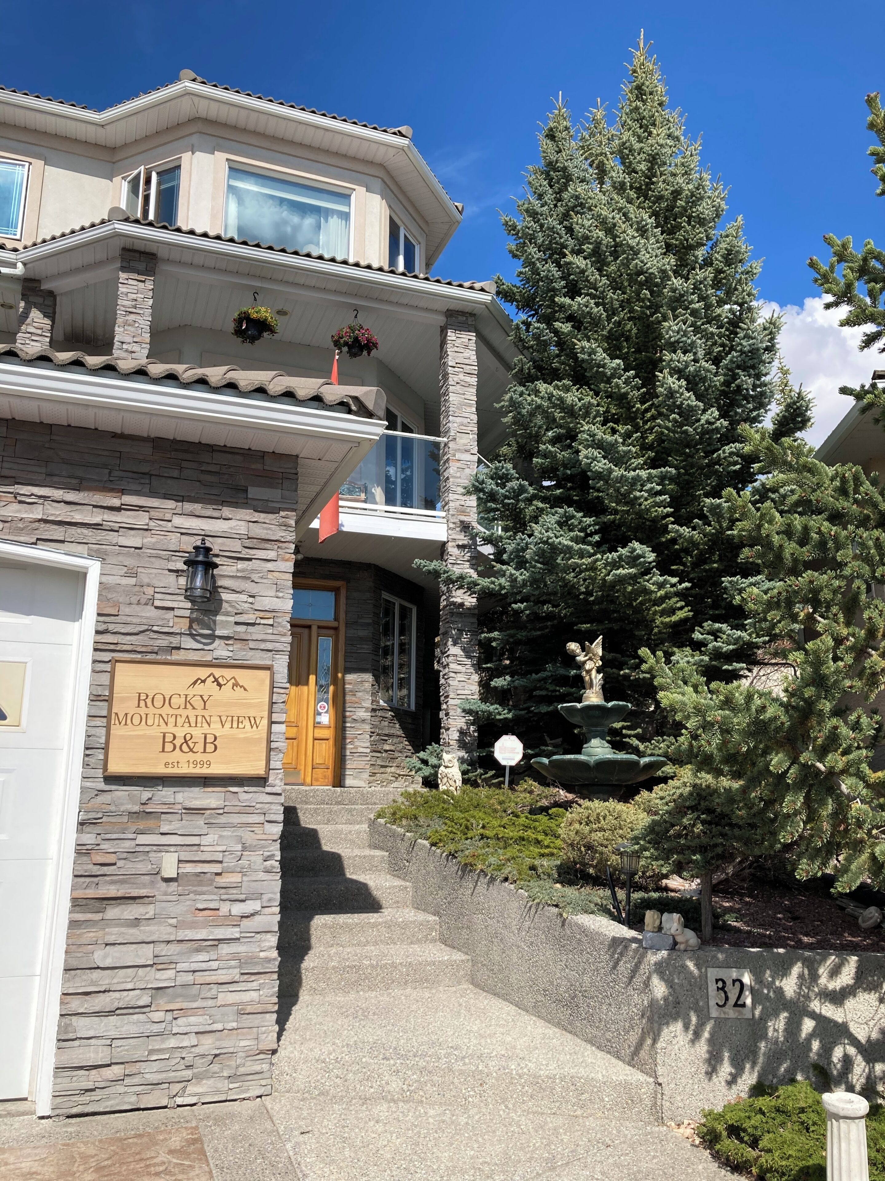 A two-story building with a stone facade, a wooden sign reading 'ROCKY MOUNTAIN VIEW B&B,' and a large evergreen tree in front.