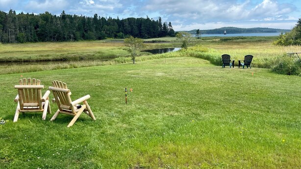 A grassy field with two wooden chairs and a table, a small tree, and a lake in the background. - Charming Newly-Renovated Waterfront Cottage in Lubec (Lubec)