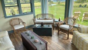 A cozy room with wicker furniture, a wooden coffee table, and large windows offering a view of a grassy field and water. - Charming Newly-Renovated Waterfront Cottage in Lubec (Lubec)