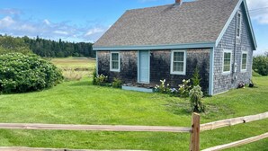 A house with a sloped roof, a front porch, and a small garden with flowers and shrubs. - Charming Newly-Renovated Waterfront Cottage in Lubec (Lubec)