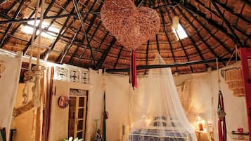 Una habitación con techo de paja, cama con dosel, un adorno colgante en forma de corazón y una estética rústica.