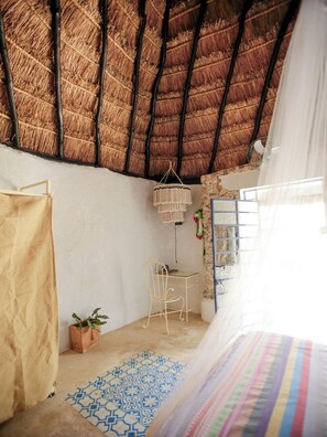 A room with a thatched roof, a bed with a striped blanket, a small table with a chair, and a hanging macrame light fixture.