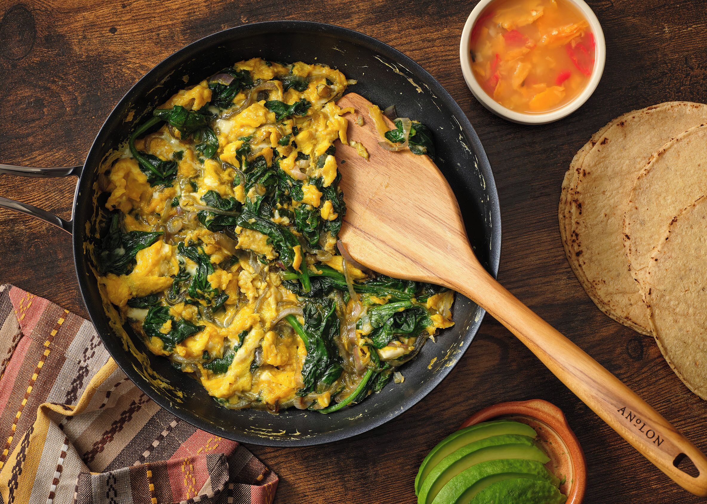 Una sartén con huevos revueltos y espinacas, una cuchara de madera, un tazón de sopa y tortillas sobre una mesa de madera.