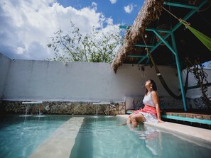 Une femme est assise au bord d’une piscine, avec une structure à toit de chaume et un mur de pierre en arrière-plan.