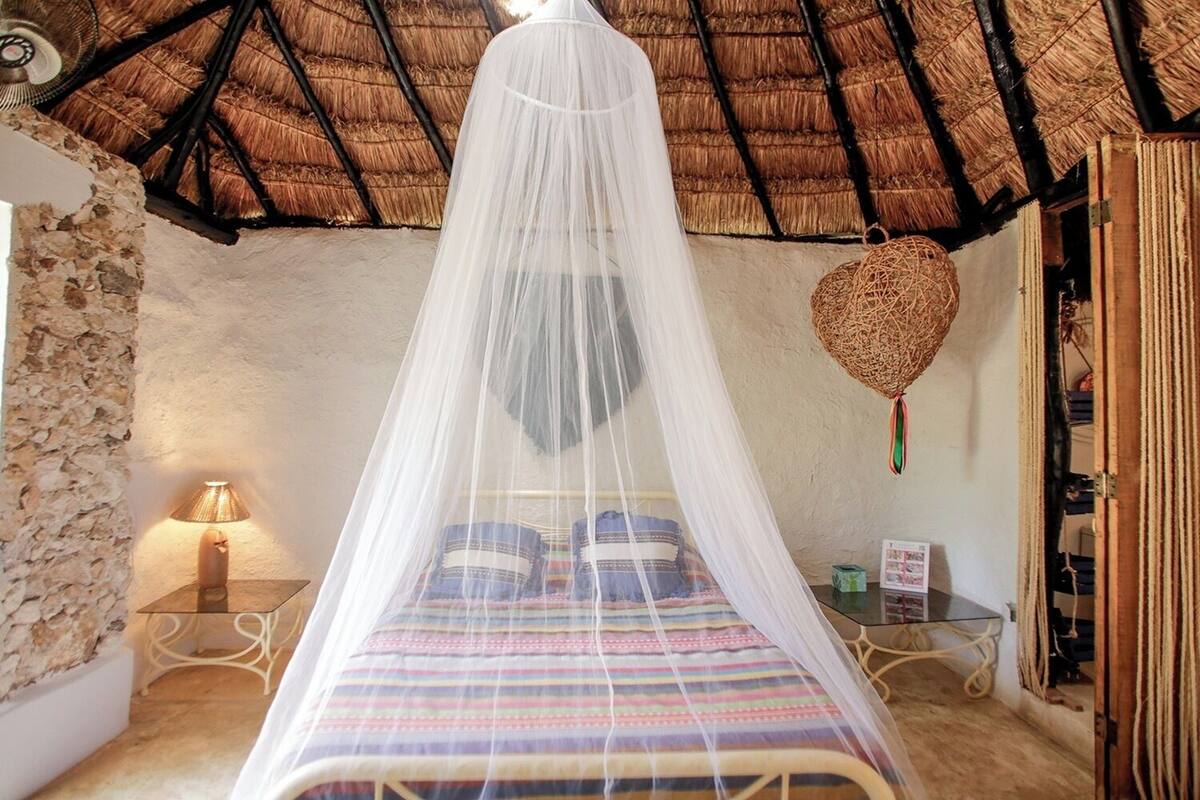 Une chambre avec un lit sous un dais, un mur en pierre, un panier tressé et une petite table avec une lampe.