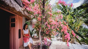 A person sitting outside a thatched-roof structure with pink flowers and tropical plants. - Hotel y Museo Aldea Maya Toktli Orígenes (Izamal)
