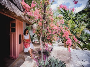 Une personne assise à l’extérieur d’une construction dont le toit est fait de chaume, entourée de fleurs roses et de plantes tropicales.