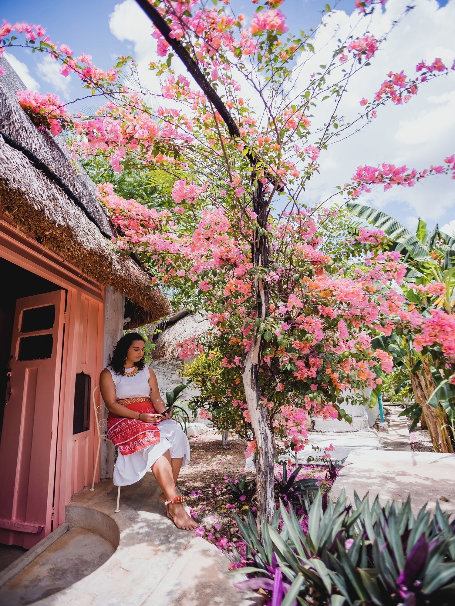 A person sitting on steps outside a building with a thatched roof, surrounded by pink flowering trees.