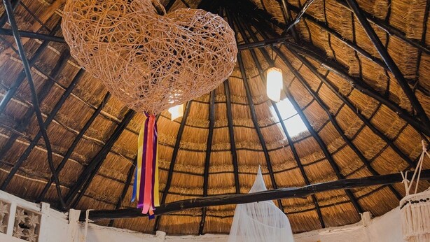 A room with a thatched roof, a bed with a blue bedspread, a hanging light fixture, and a white wall. - Hotel y Museo Aldea Maya Toktli Orígenes (Izamal)