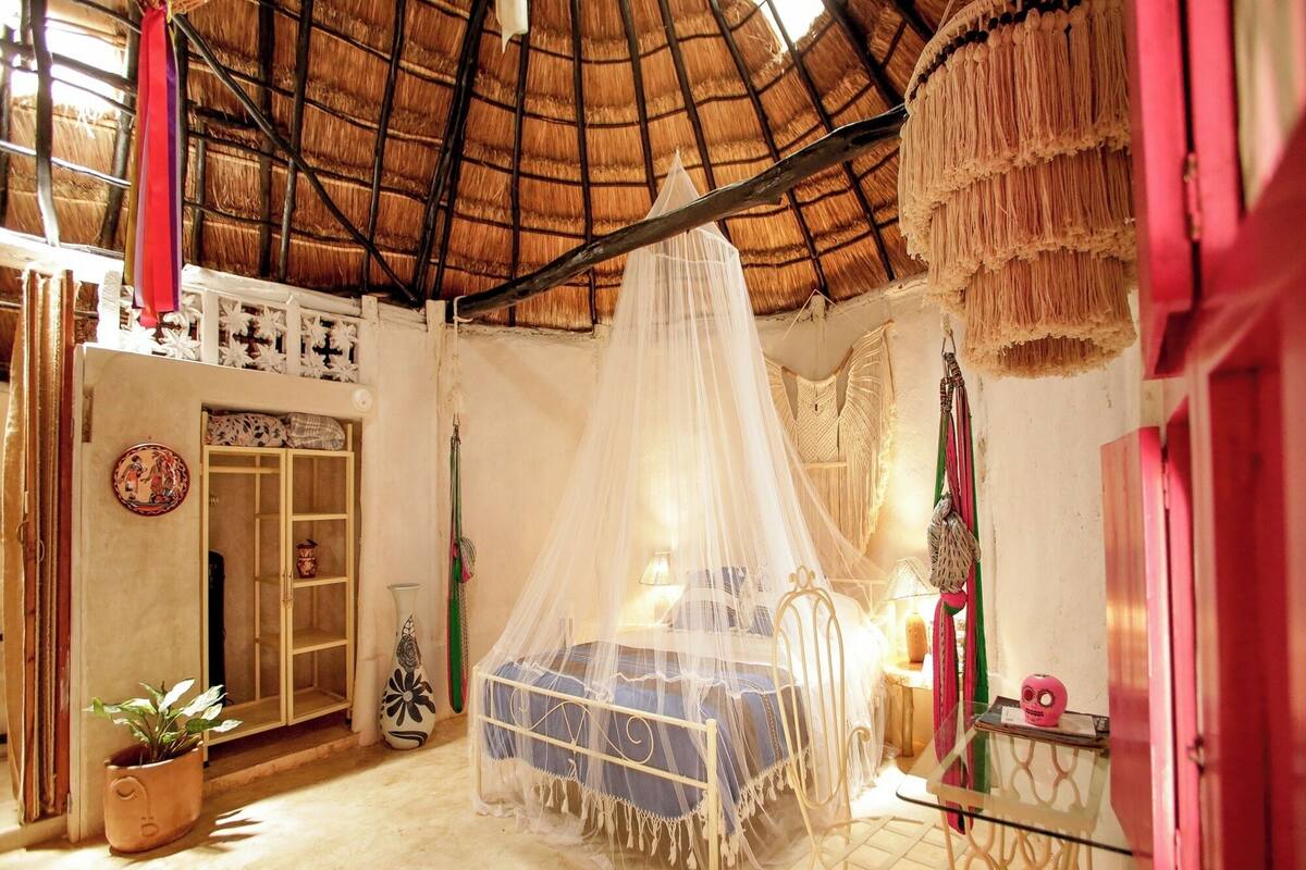 Une chambre avec un lit sous un dais, une petite table, une chaise, une plante en pot et une décoration murale.