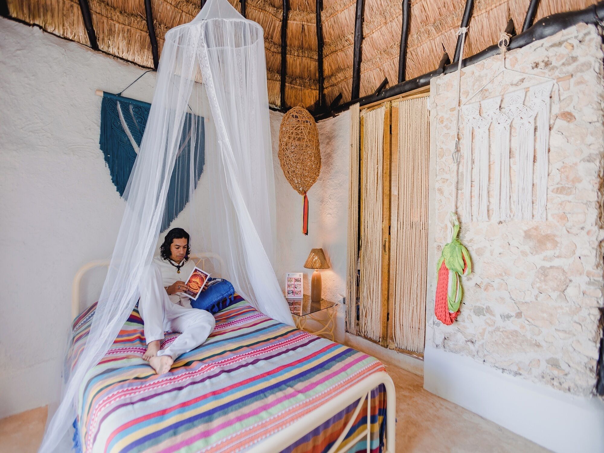 A room with a bed under a white canopy, a stone wall, and a woven wall hanging.