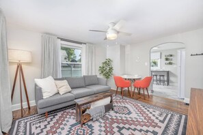 A modern living room with a grey sofa, a coffee table, and a patterned rug. There is a dining area with a small table and orange chairs.