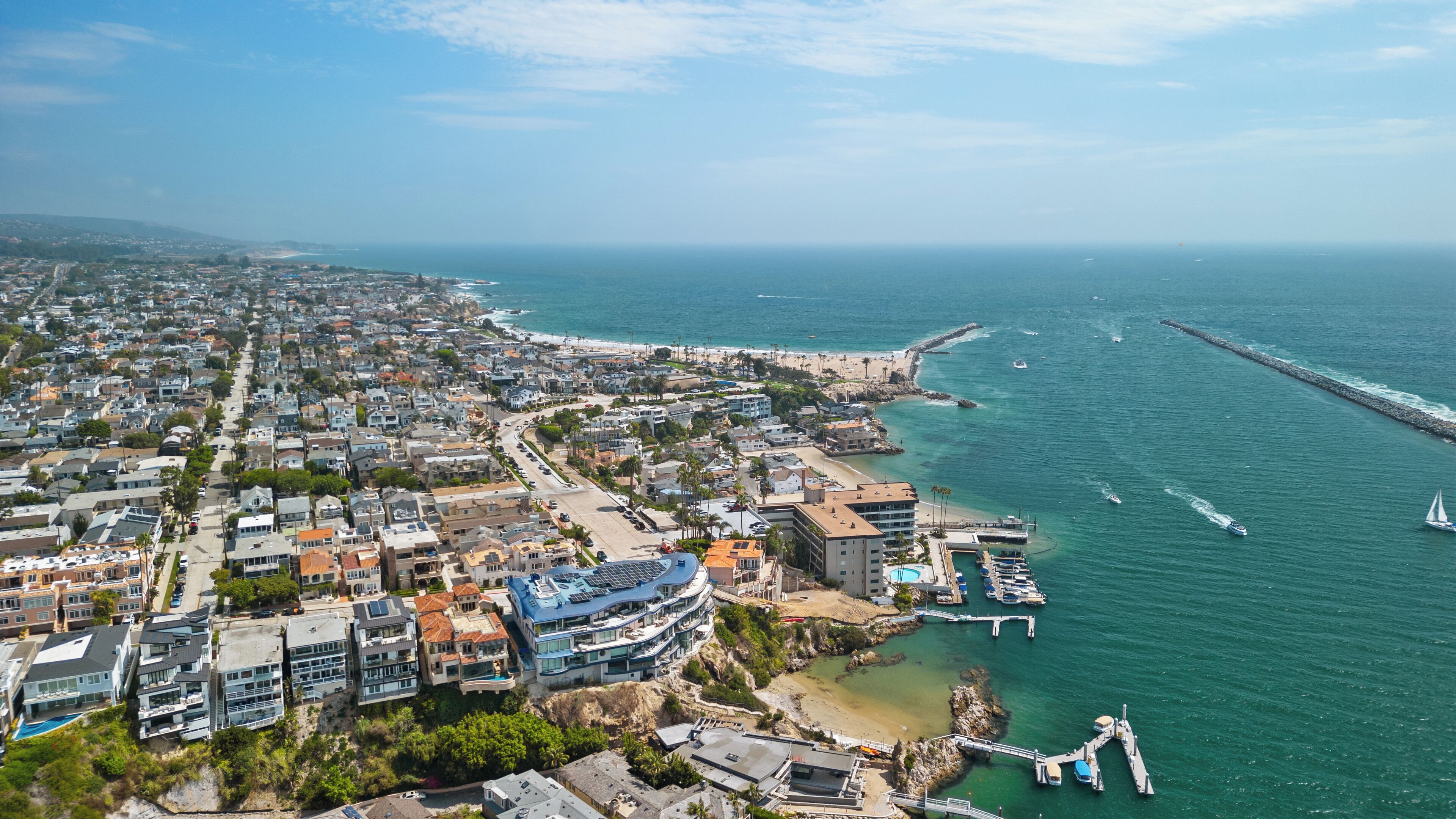 An aerial view of a coastal town with residential buildings, a marina, and a pier extending into the sea.