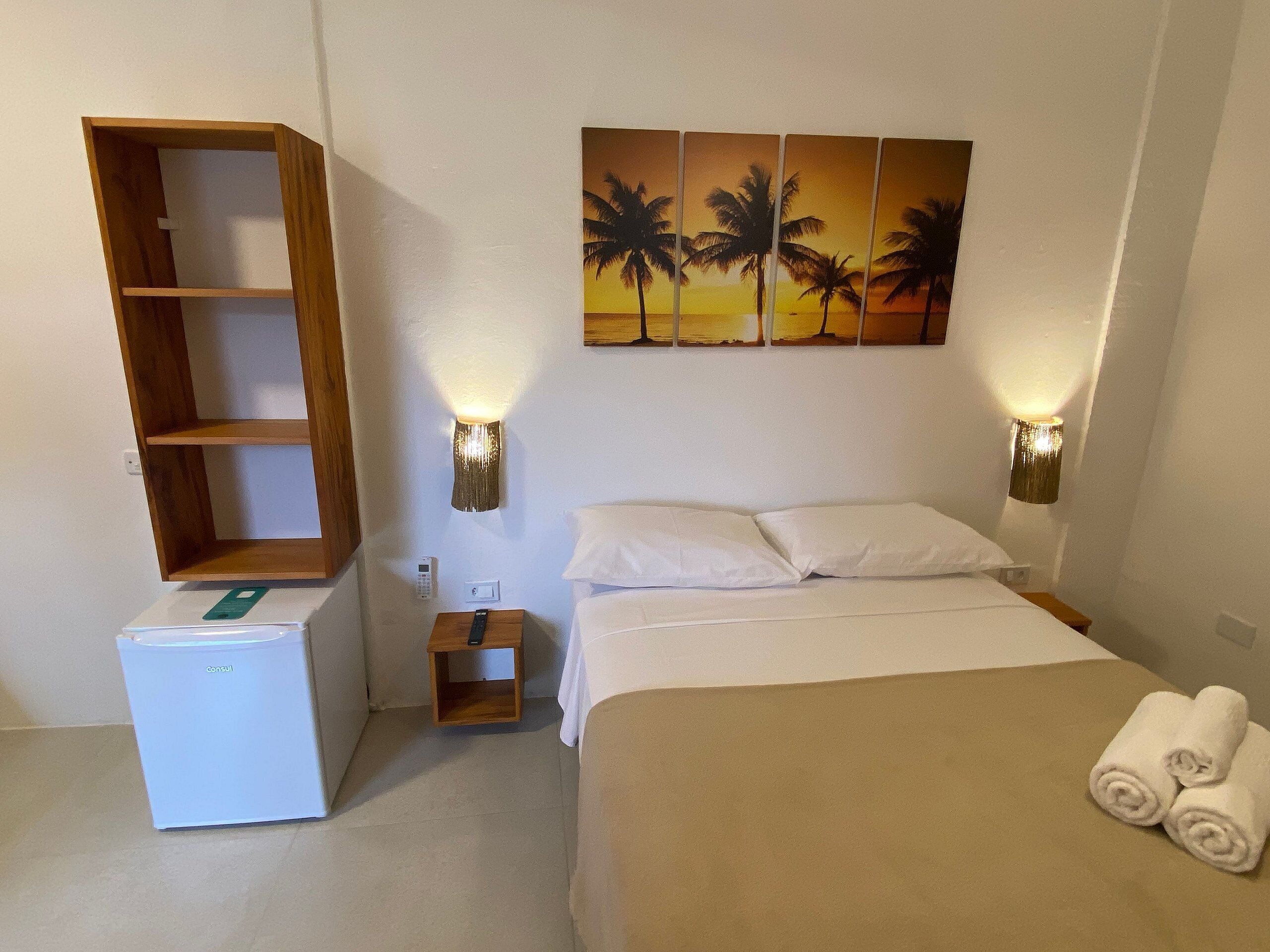 A hotel room with a bed, a bedside table, a small wooden shelf, a bedside lamp, and a wall-mounted painting of palm trees.