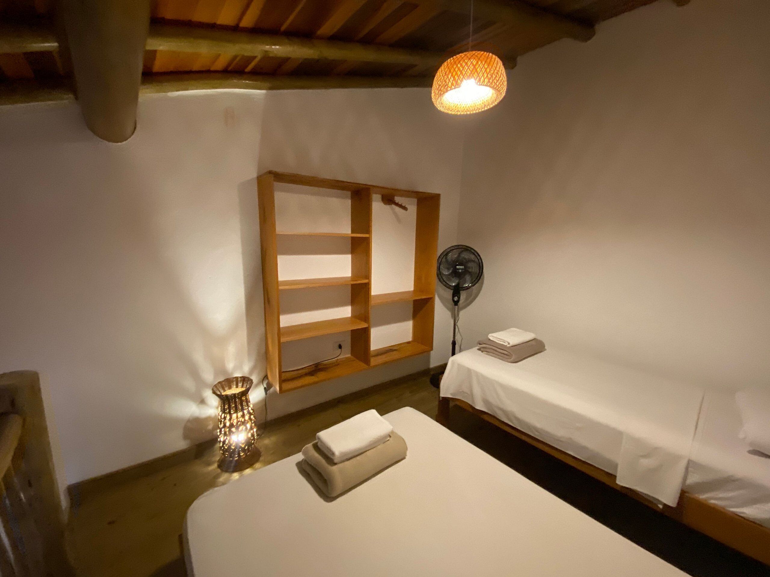 A small, cozy room with a single bed, a bedside table, a wall-mounted shelf, a floor lamp, and a ceiling fan.
