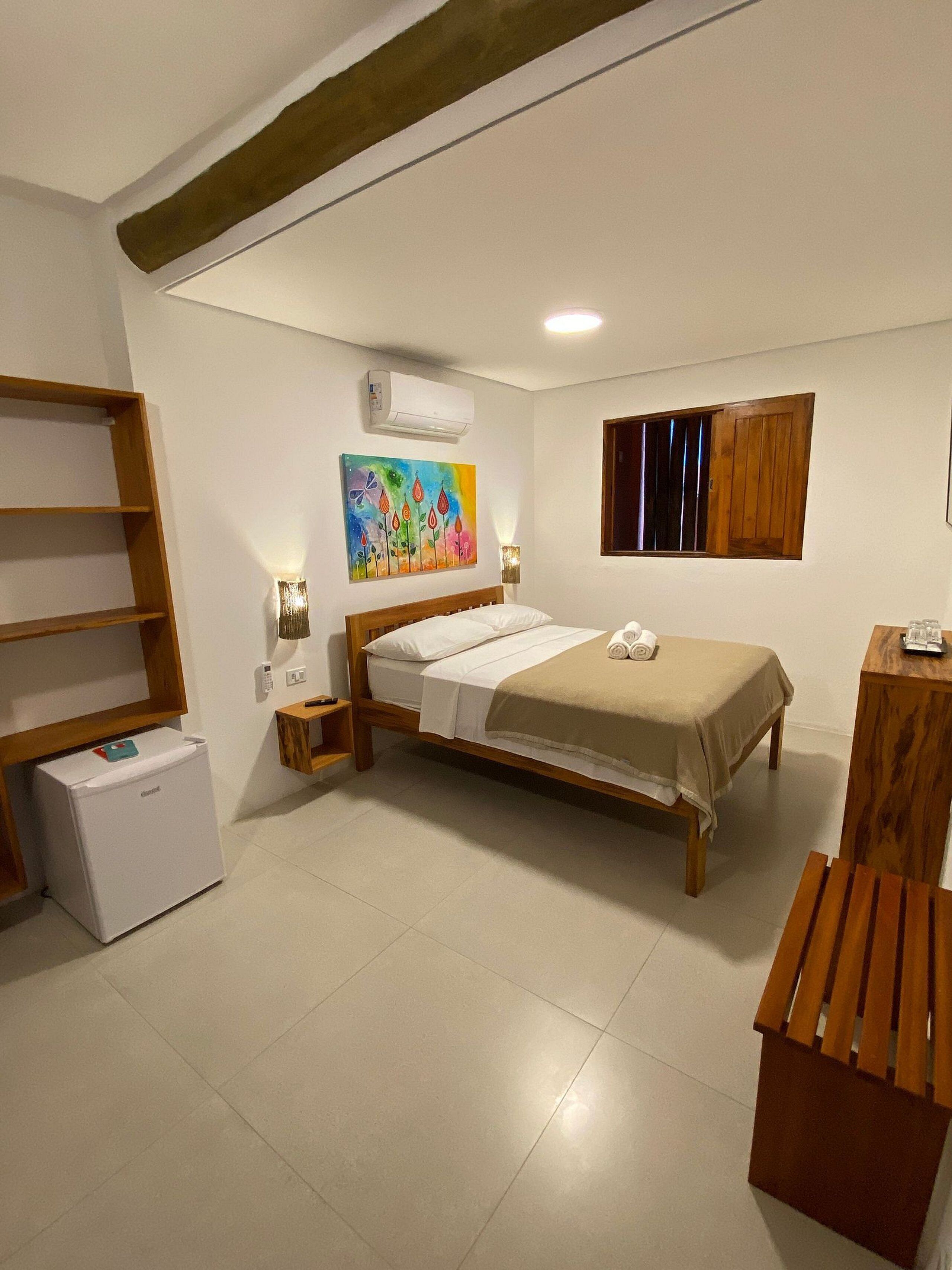 A bedroom with a bed, a small bedside table, a nightstand, a wooden bench, a wardrobe, a wall-mounted air conditioner, a colorful painting, and a window with wooden shutters.