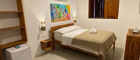 A bedroom with a bed, a small bedside table, a nightstand, a wooden bench, a wardrobe, a wall-mounted air conditioner, a colorful painting, and a window with wooden shutters.