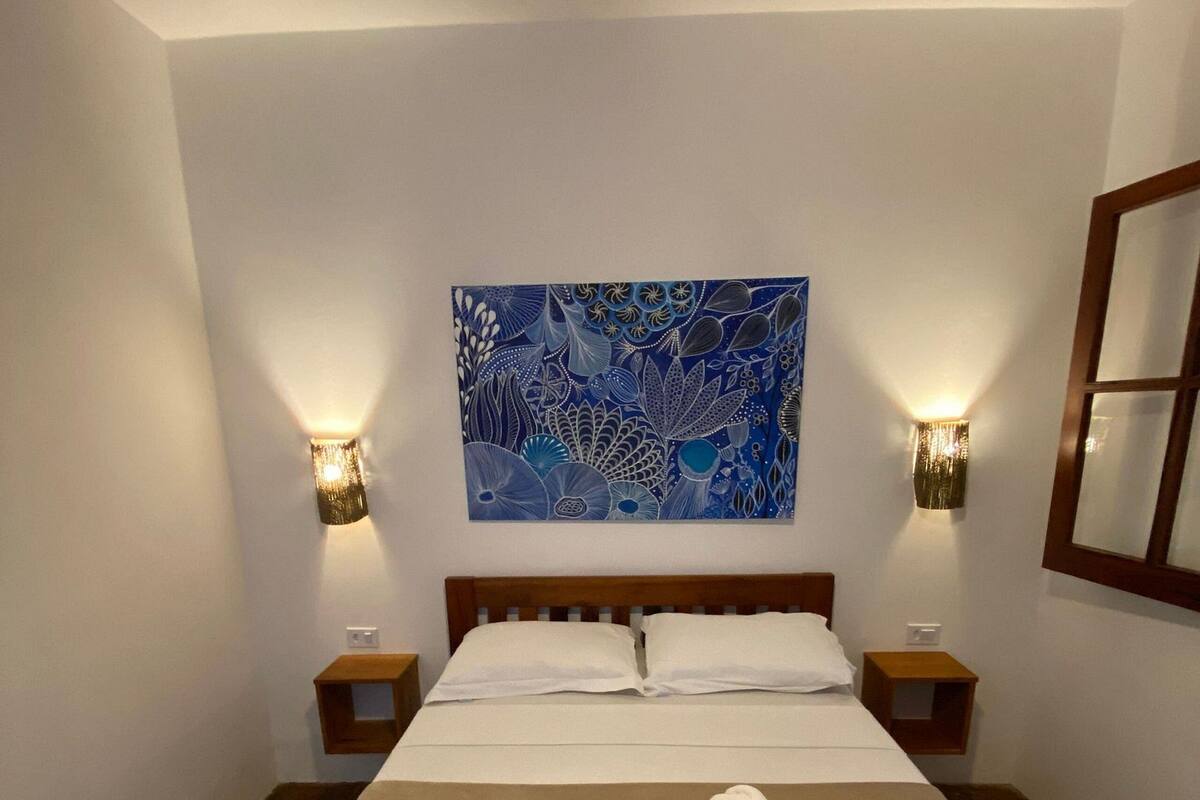 A neatly made bed with white linens and three rolled towels on the bed. There is a large blue abstract painting on the wall above the bed.