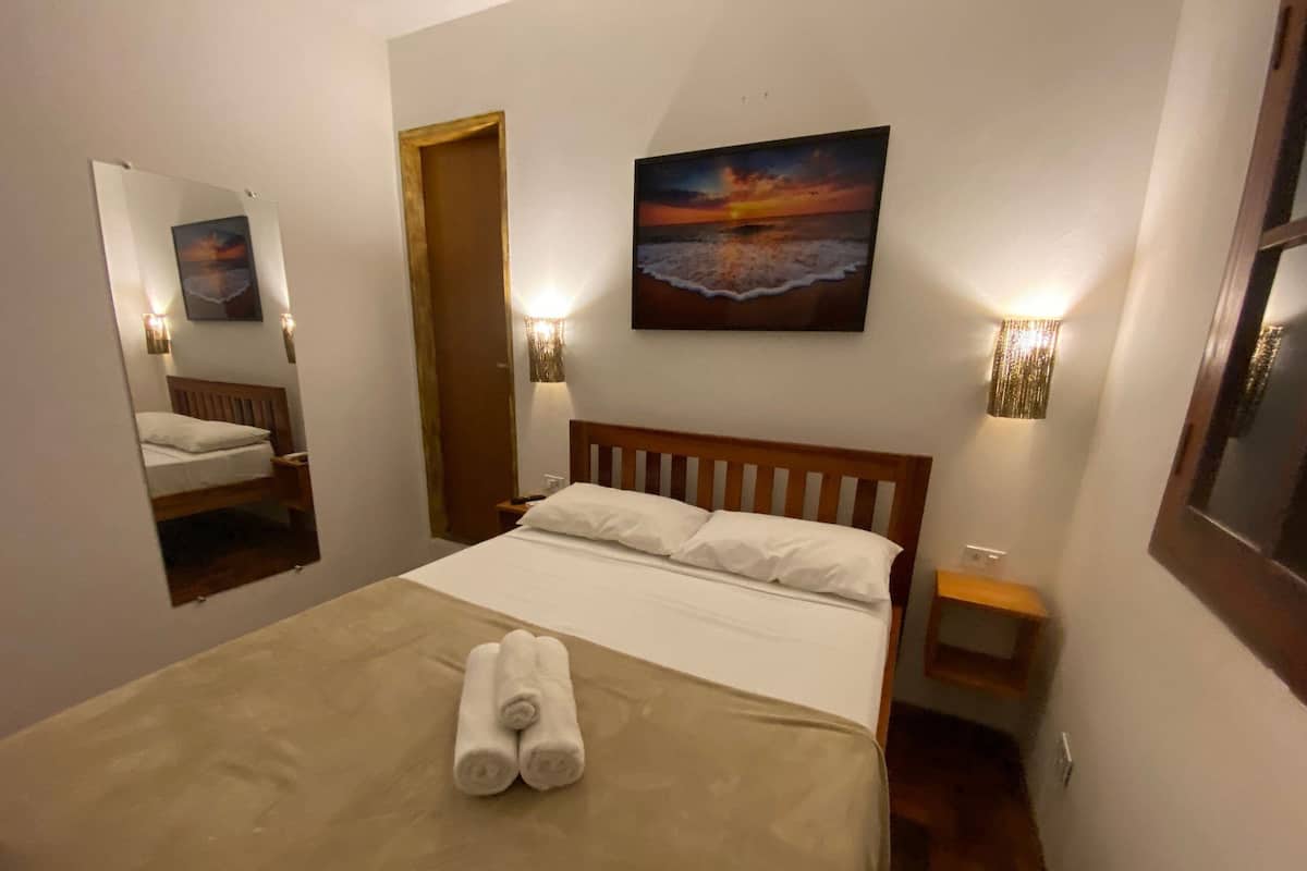A neatly made bed with white linens and two rolled towels, a painting of a sunset over the ocean on the wall, and two wall-mounted lamps on either side of the bed.