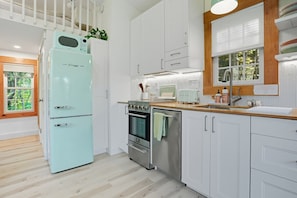 A modern kitchen with white cabinetry, a stainless steel oven, a light blue refrigerator, and a window with a view of greenery. - Mountain-side modern tiny cabin with fire pit (West Windsor)