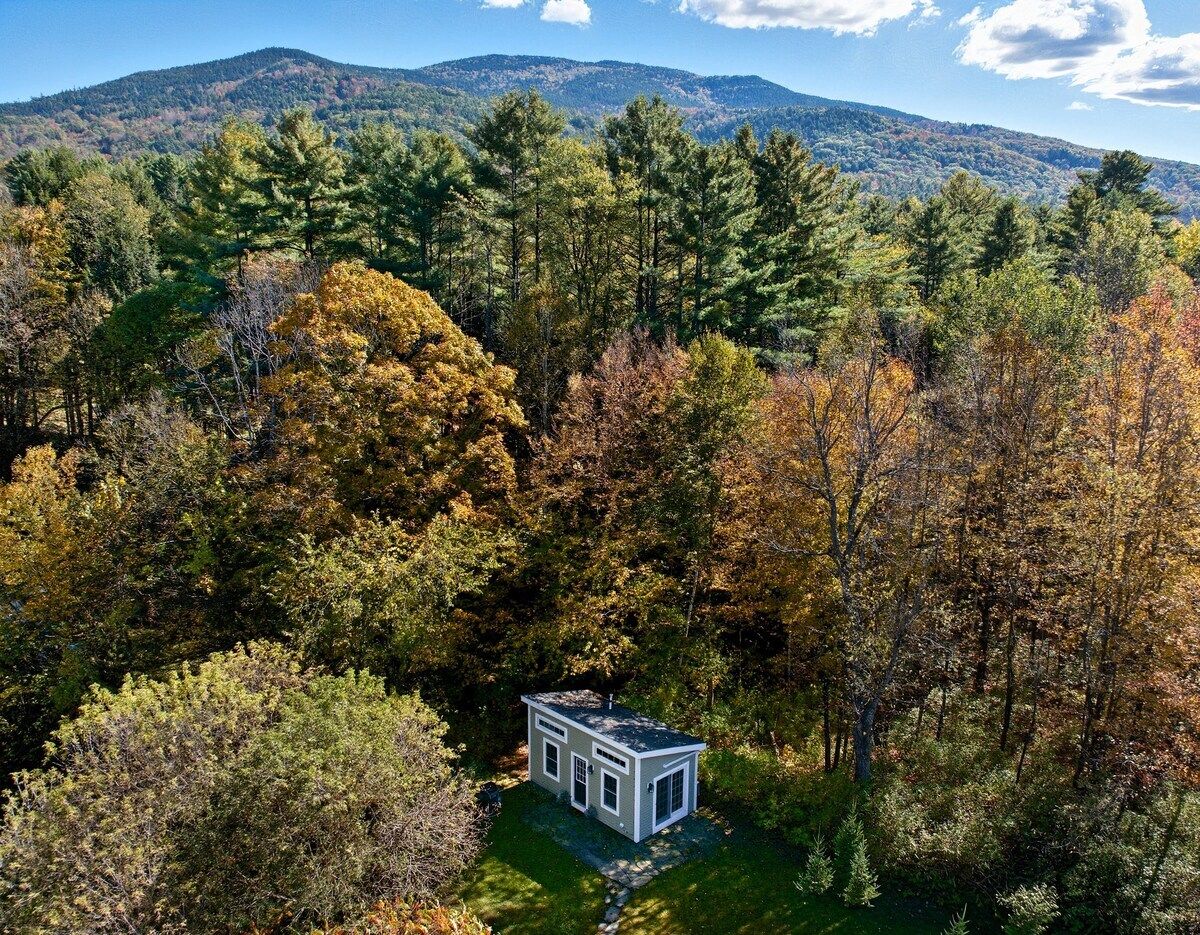 A small, white cabin surrounded by dense forest with a mountain in the background.