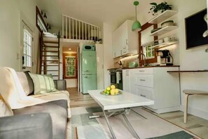 A compact living space with a sofa, a dining table with fruit, a kitchenette, and a staircase.