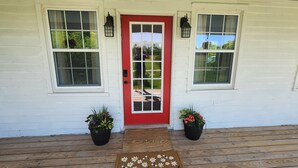 Une maison blanche avec une porte rouge, deux plantes en pot et un porche en bois.