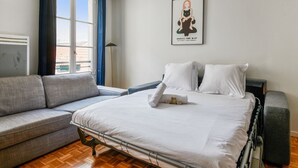 A modern hotel room with a large bed, a grey sofa, and a window with curtains. - Appartement Garnet - Welkeys (Paris)
