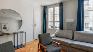 A modern living room with a wooden floor, a grey sofa, a wooden chair, and a round mirror on the wall. - Appartement Garnet - Welkeys (Paris)
