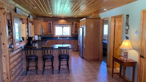 A log cabin-style kitchen with wooden cabinets, a central island with bar stools, and a dining table. - Brevort Log Cabin, Michigan's UP (Moran)