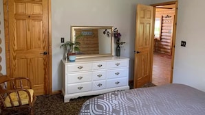A bedroom with a wooden ceiling, a white dresser with drawers, a bed with patterned bedding, and a wooden door. - Brevort Log Cabin, Michigan's UP (Moran)
