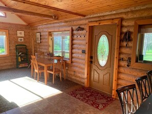 Interior de una cabaña de troncos con comedor, una puerta con vidriera y una estantería.