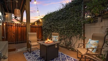 An outdoor patio with a fire pit, two chairs, a patterned rug, and a wooden fence.