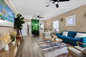 Een ruime woonkamer met een blauwe bank, houten stoelen, een salontafel en een grote plant. Een plafondventilator is zichtbaar, en er is een groene muur met een raam.