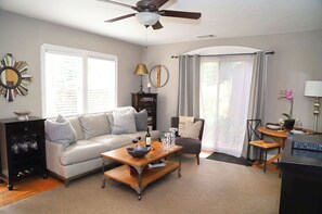 A living room with a sofa, coffee table, and a chair. There is a ceiling fan, a window with curtains, and a bookshelf. - The Downtown Napa Getaway (Napa)