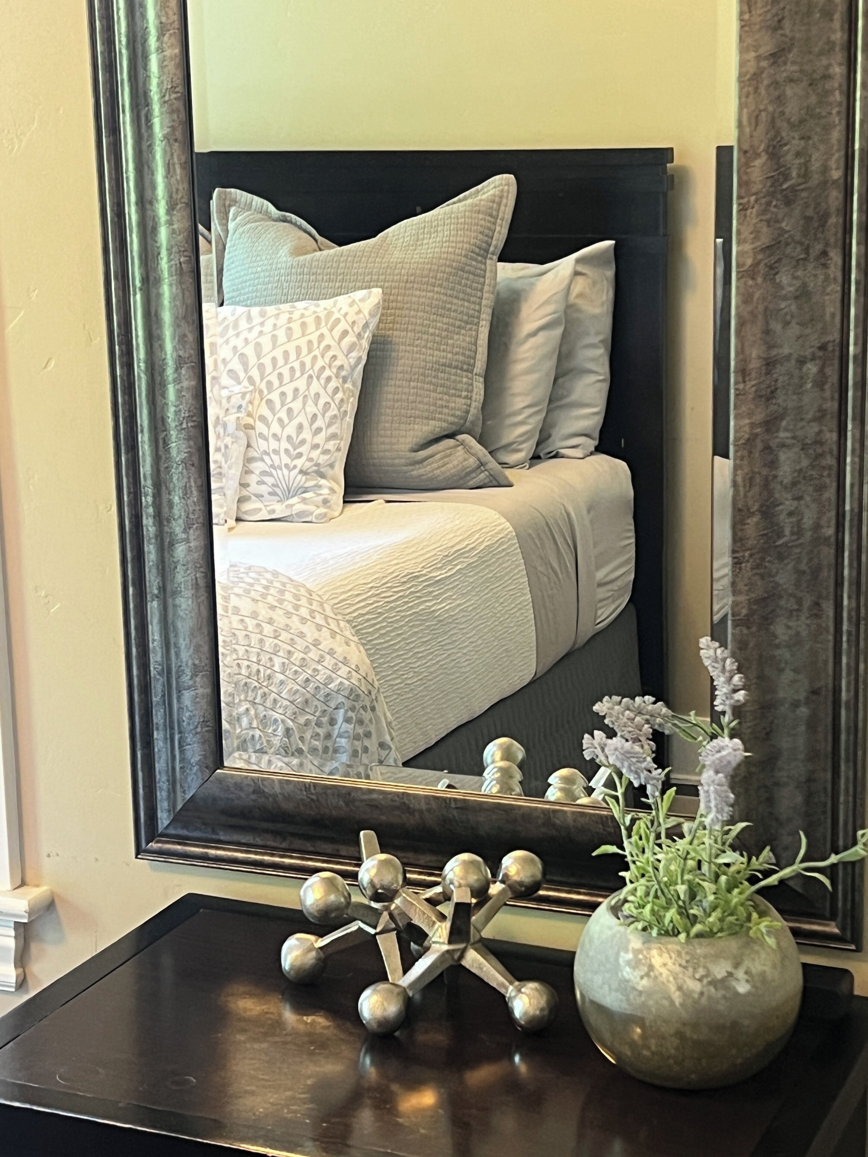 Un dormitorio con una cama, almohadas, un espejo, un jarrón con flores y una escultura metálica decorativa sobre una mesa.