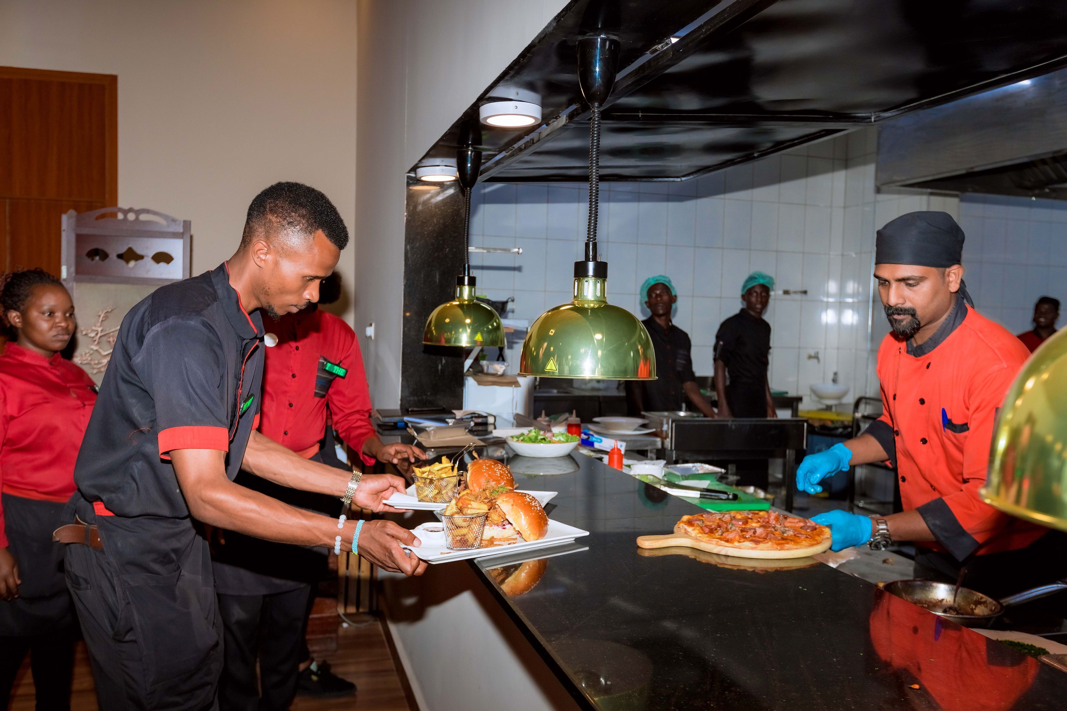 A kitchen area with chefs preparing food, a pizza on the counter, and various kitchen utensils and ingredients.
