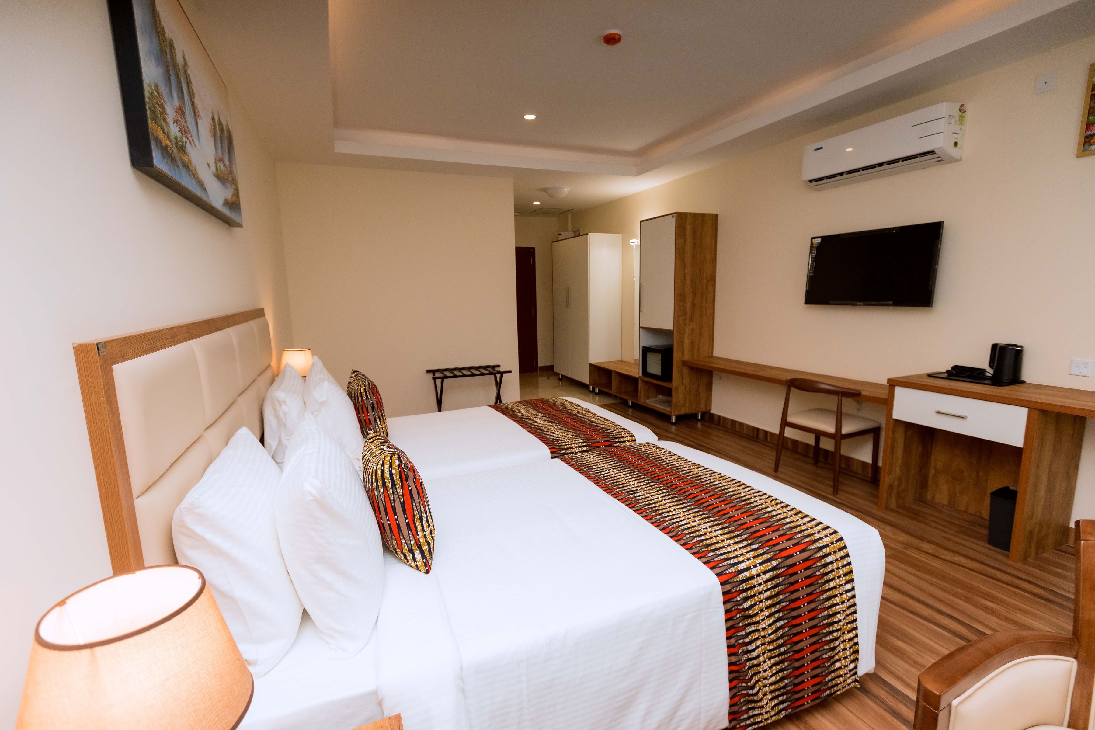 A hotel room with a bed, bedside table, lamp, desk, chair, TV, and air conditioning unit.