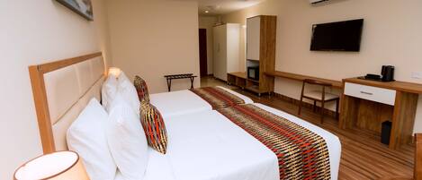 A hotel room with a bed, bedside table, lamp, desk, chair, TV, and air conditioning unit.